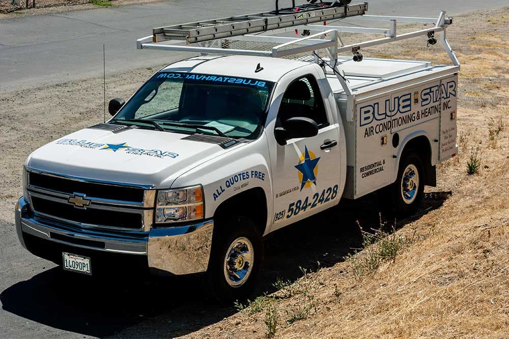 Zena, a white Blue Star Chevy Silverado 3500HD utility truck with a ladder rack, parked beside dry grass