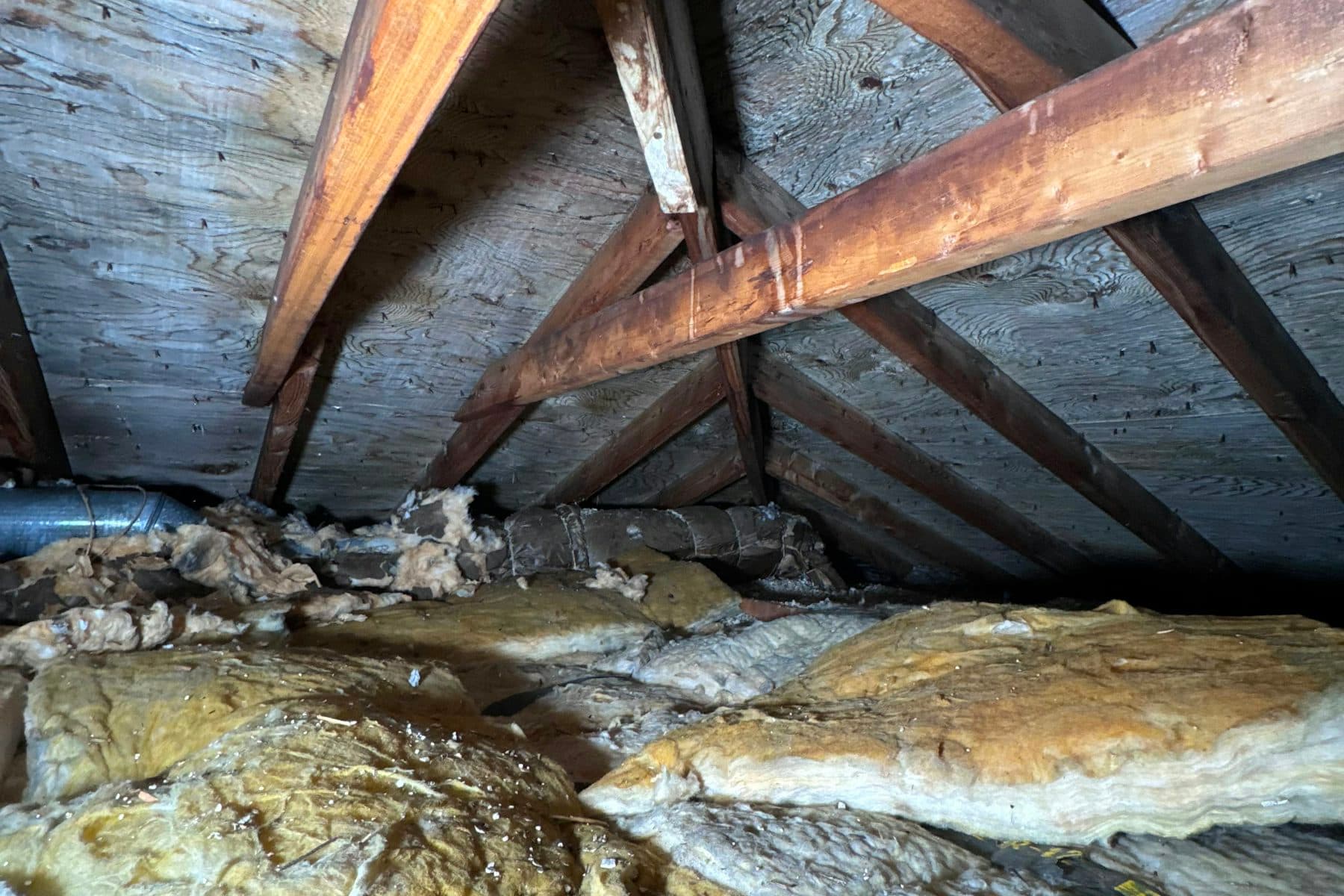 Old yellow fiberglass batts and a metal duct run beneath wooden rafters in a dim attic