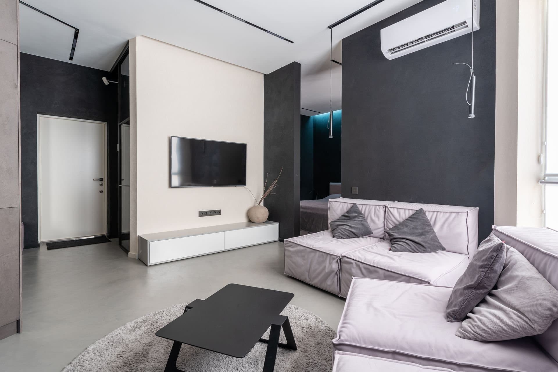 Wall-mounted ductless mini-split head above a modern living room with grey sofas and a TV