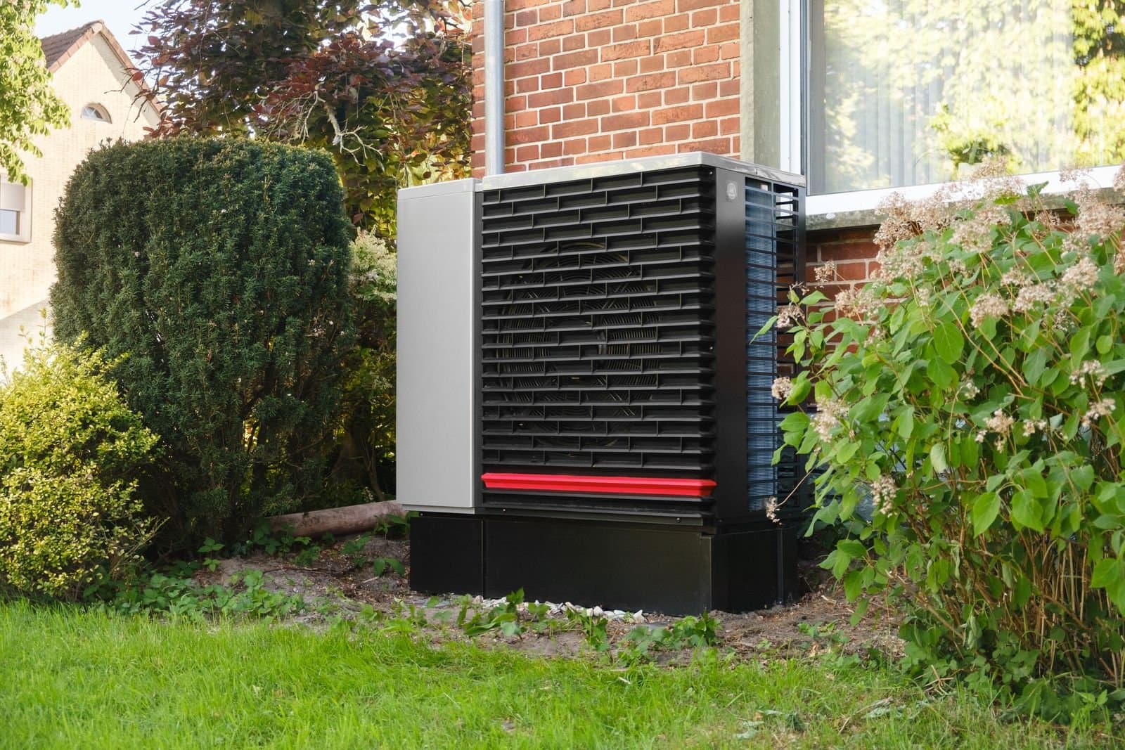 Outdoor heat pump unit on a lawn beside a brick home, surrounded by garden shrubs