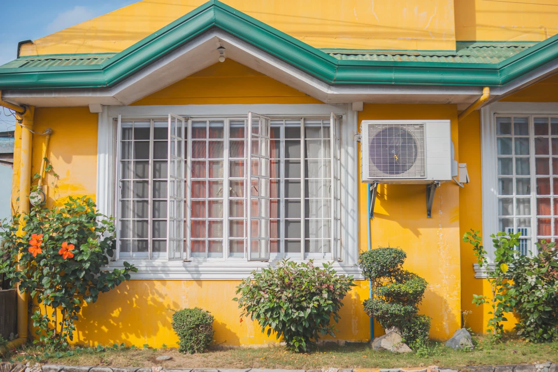 Mini-split outdoor unit mounted on the sunny yellow wall of a single-story house with flowering shrubs