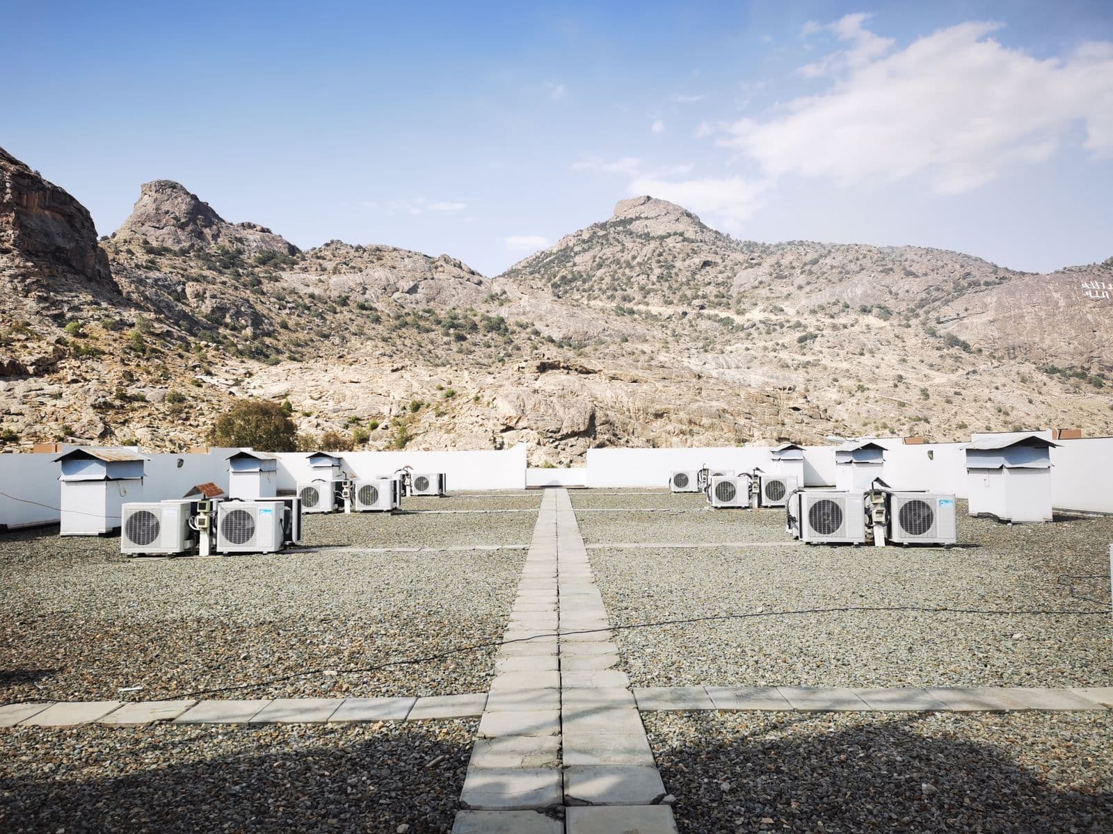 Rows of air conditioning condensers on a flat gravel rooftop with dry rocky mountains behind