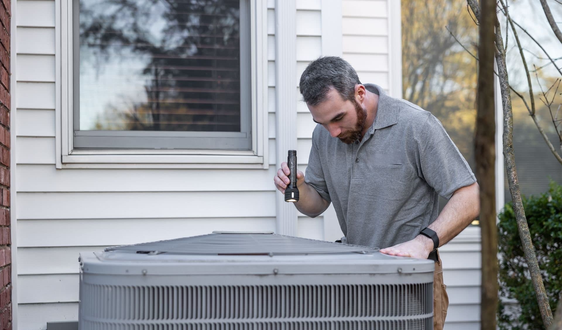 Technician with a flashlight inspecting the top of an outdoor AC condenser beside a white-sided home