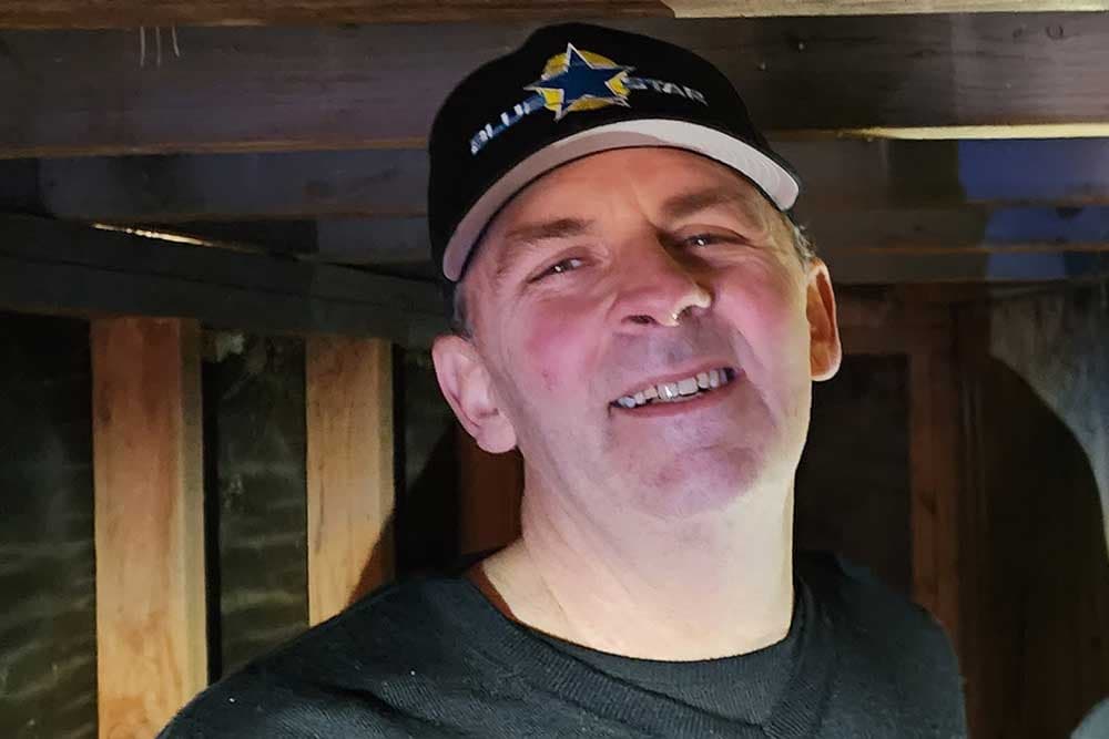 Dennis Carter of Blue Star smiling in a black Blue Star cap under exposed floor joists