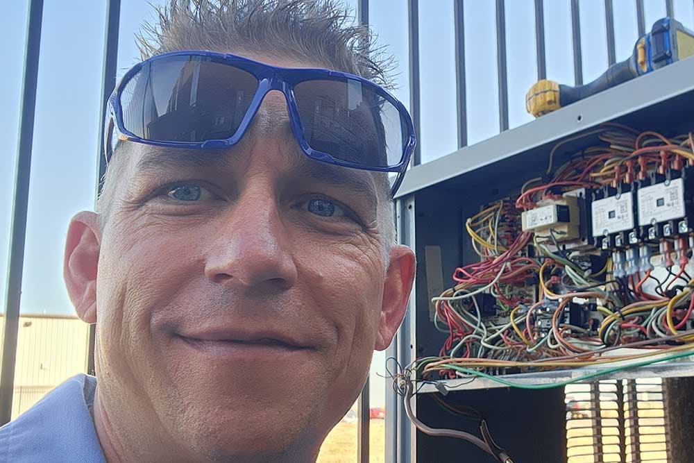 Jeff Scalier, owner of Blue Star, smiling in blue sunglasses beside the open control panel of a rooftop HVAC unit