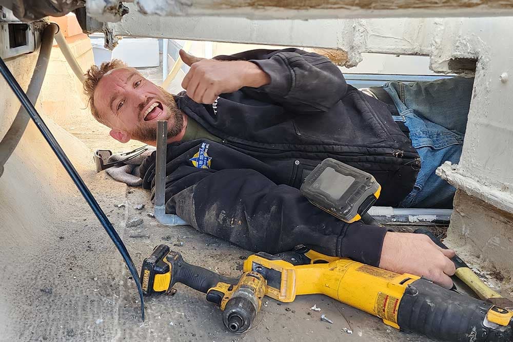 Travis of Blue Star lying under a rooftop unit giving a thumbs up, with power tools beside him