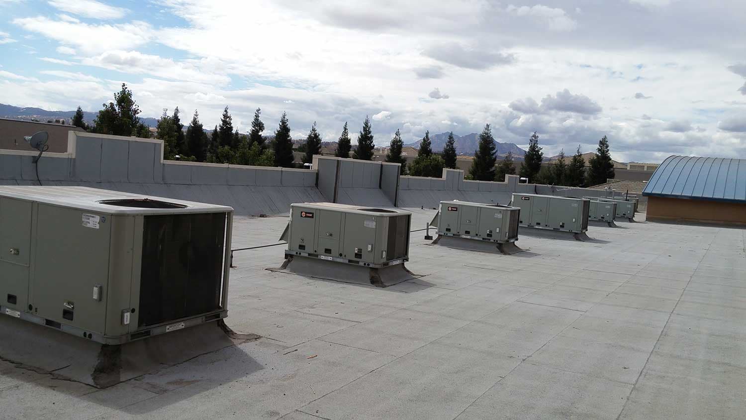 Row of packaged rooftop HVAC units on a flat commercial roof, with trees and hills on the horizon