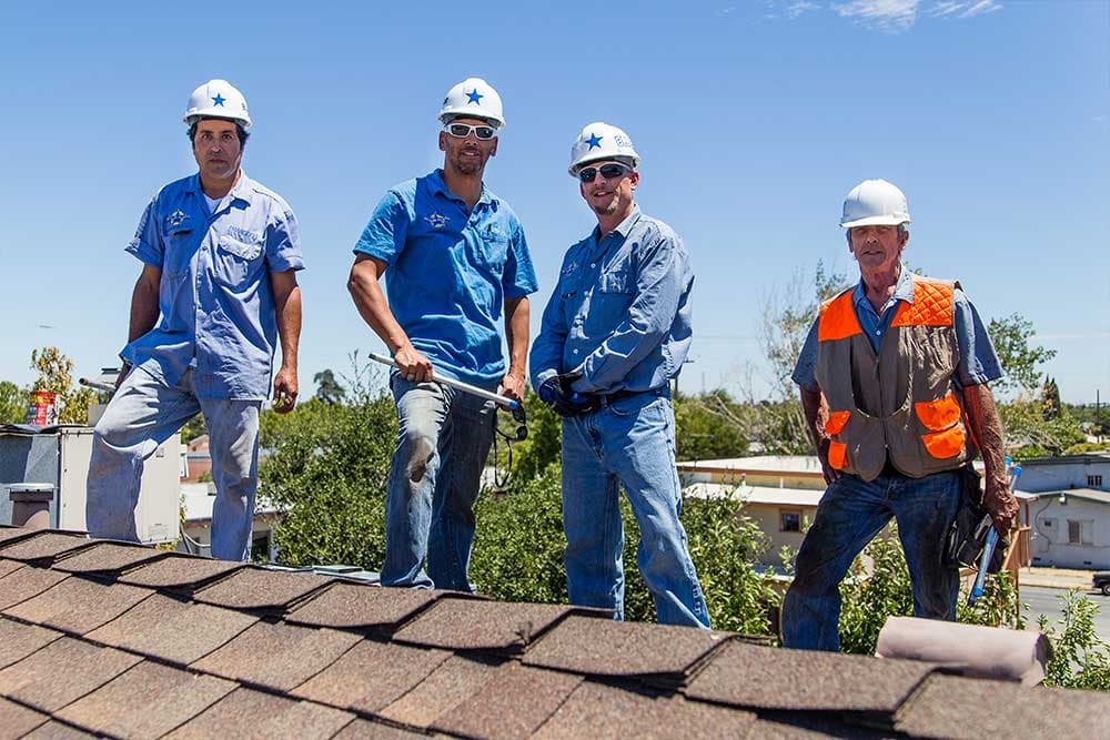 Four Blue Star technicians in hard hats standing on a shingle roof above a residential neighborhood