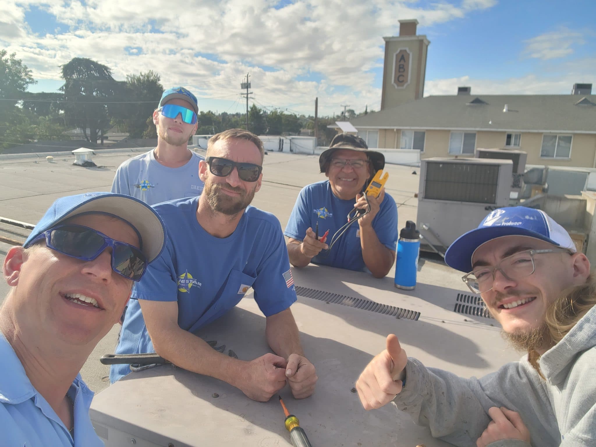 Five smiling Blue Star technicians taking a selfie on a commercial roof beside an HVAC unit