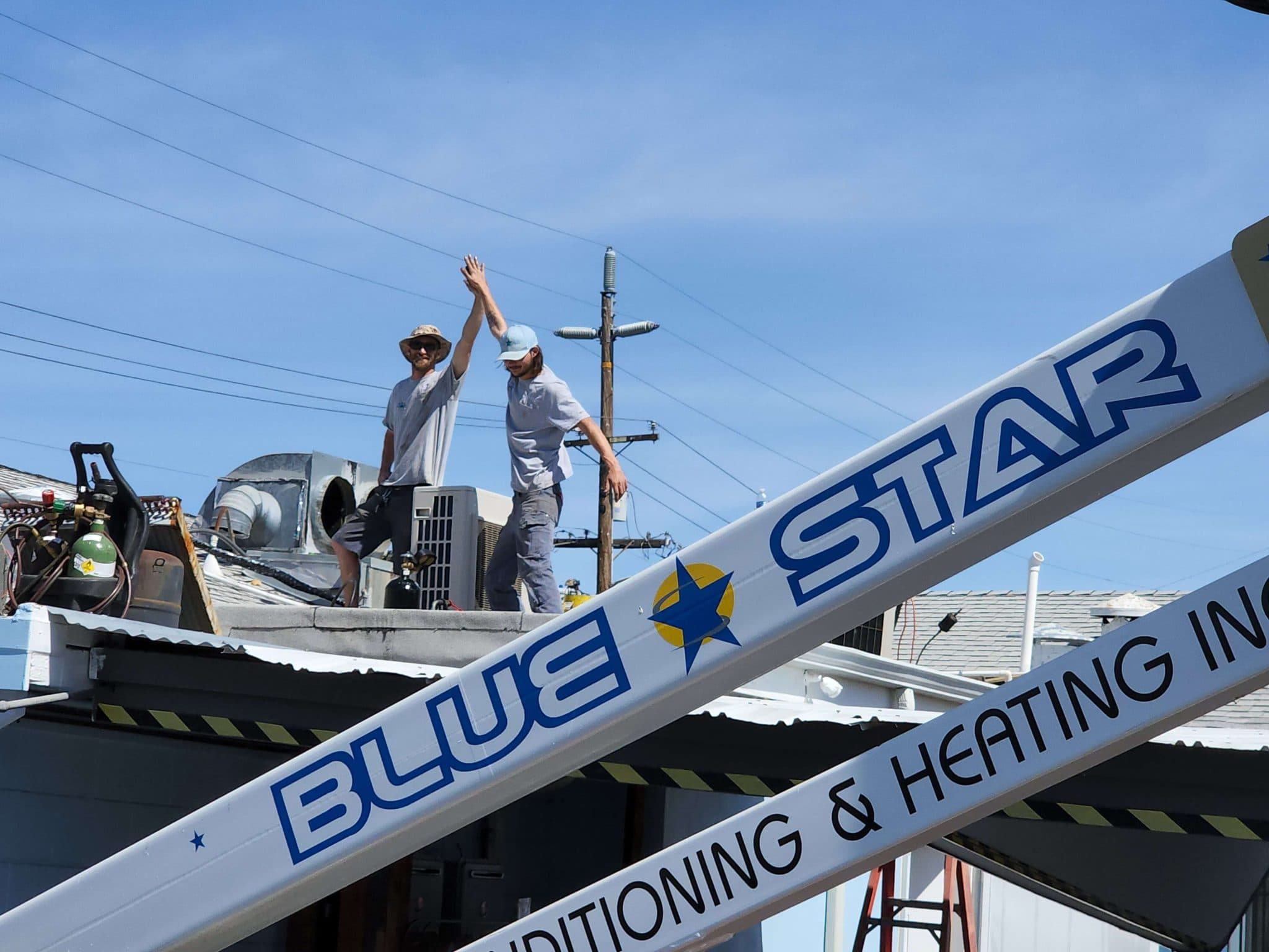 Two Blue Star technicians high-fiving on a commercial roof beyond the company's Blue Star-lettered lift boom