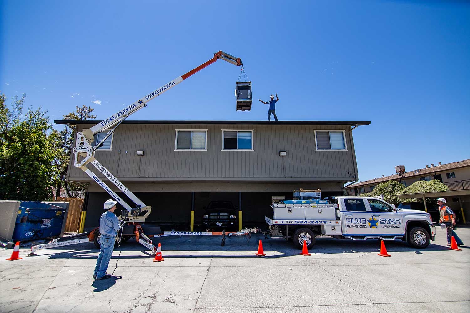 Blue Star's lift raising an HVAC unit onto a two-story apartment roof while the crew works from the coned-off parking lot below