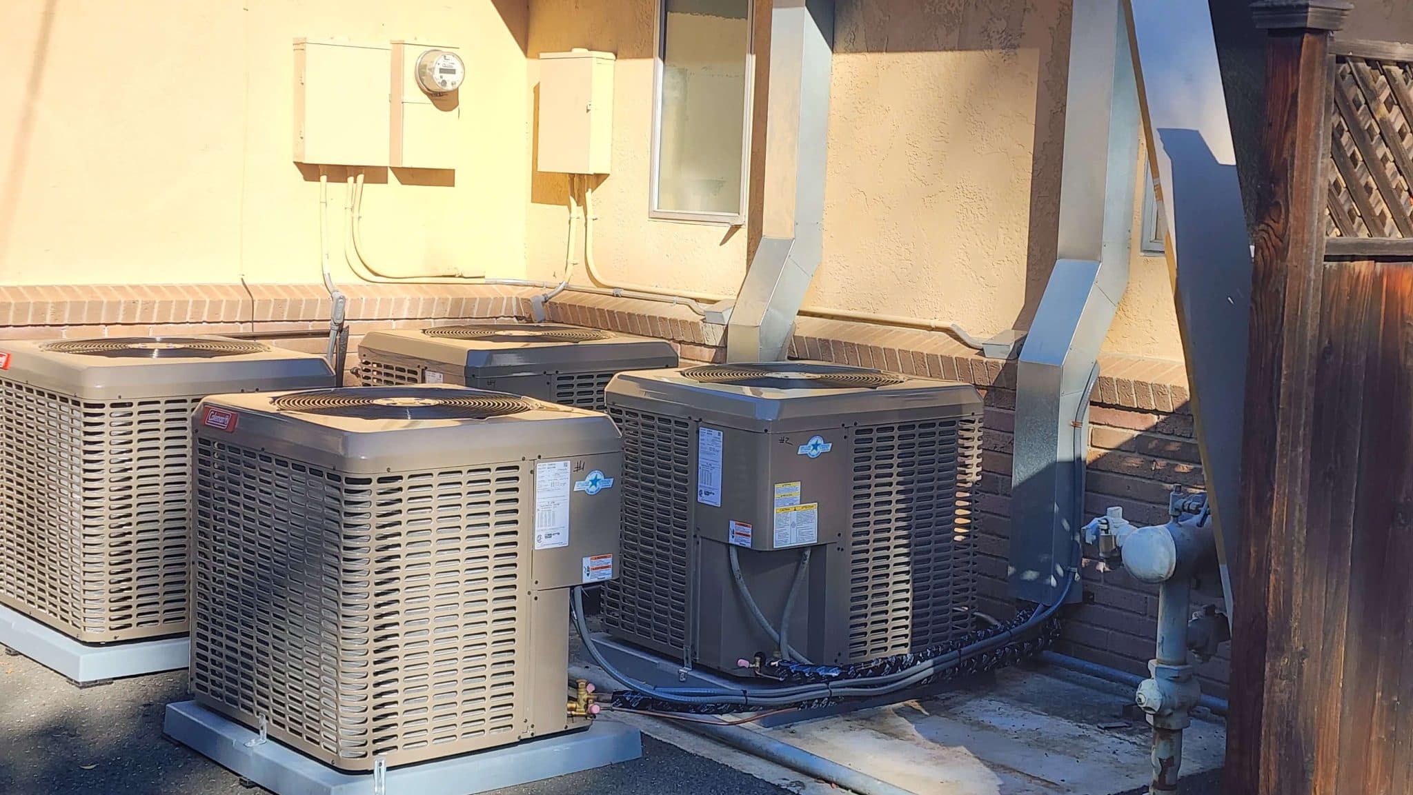 Three new outdoor AC condensers on pads beside a stucco building, with new refrigerant lines and disconnect boxes on the wall
