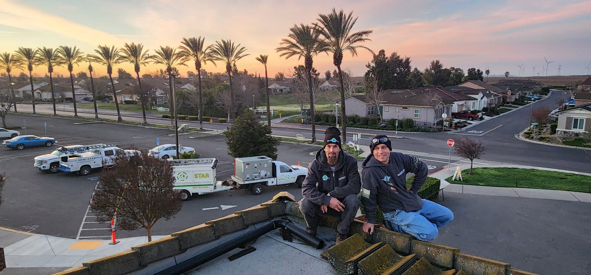 Two Blue Star technicians sitting on a roof edge at dusk above a palm-lined street and parked Blue Star trucks
