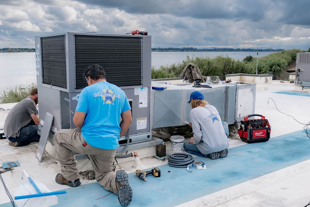 Three Blue Star technicians connecting a new packaged rooftop unit on a waterfront commercial roof