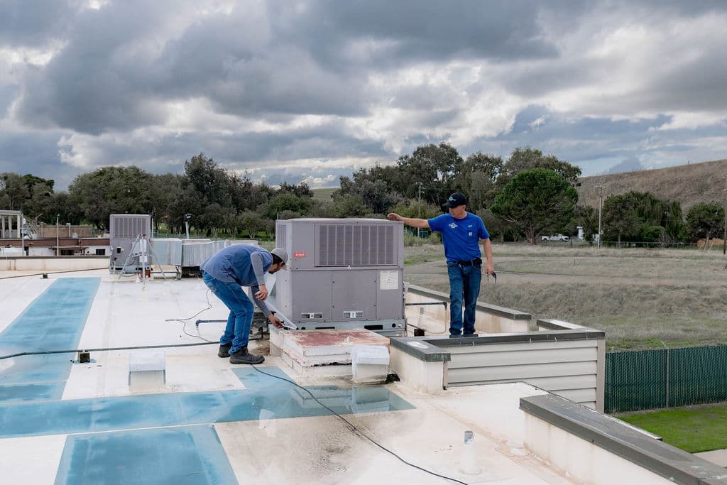 Two Blue Star technicians servicing a packaged rooftop unit on a commercial roof with green hills behind