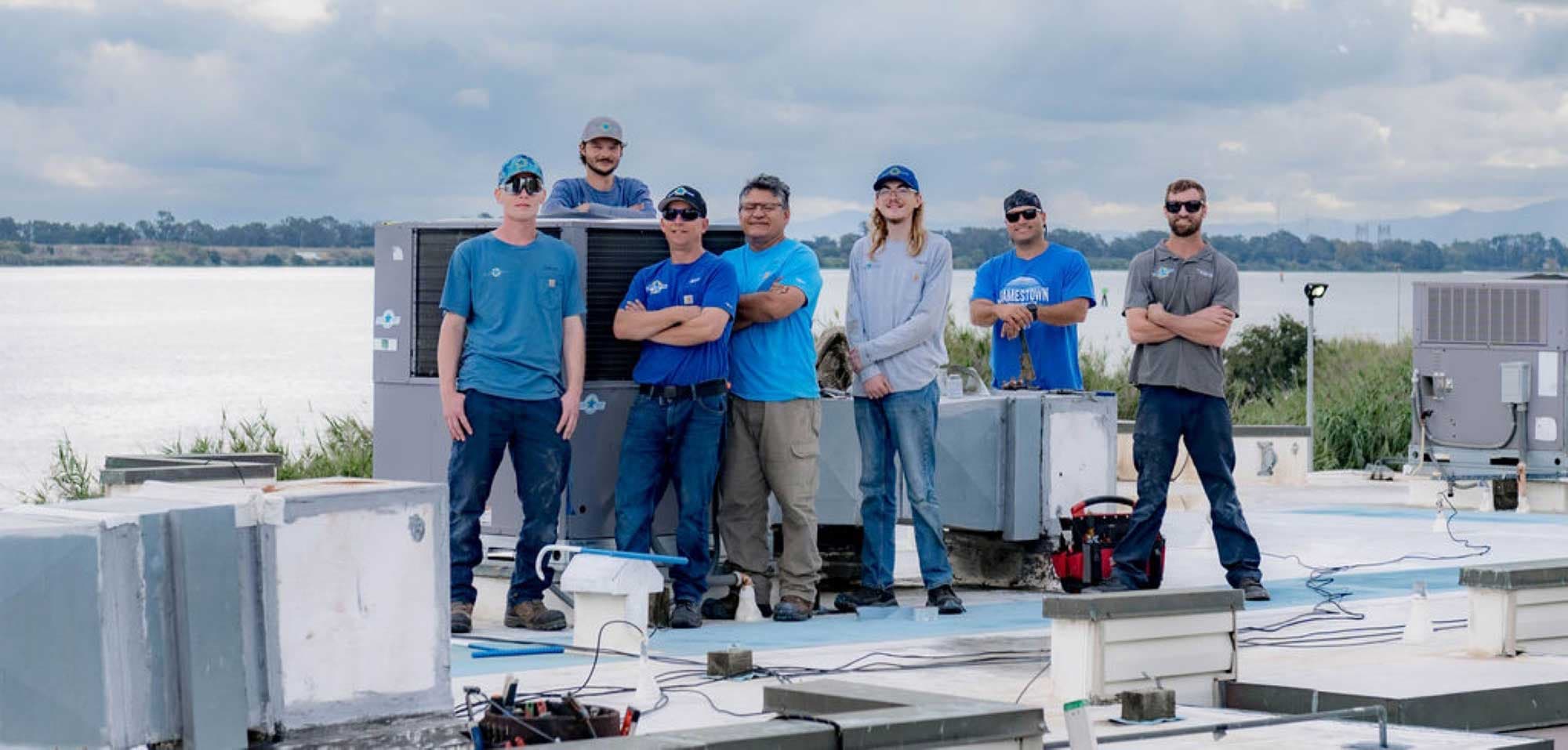 Seven Blue Star team members posing beside a new packaged rooftop unit on a flat roof overlooking the water