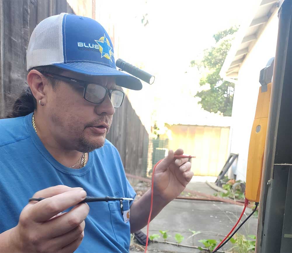 Blue Star technician holding multimeter test leads while diagnosing an outdoor AC unit in a side yard