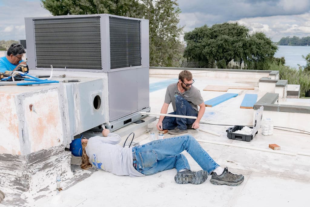 Blue Star technician lying on the roof to work under a rooftop unit's duct curb while a colleague measures pipe