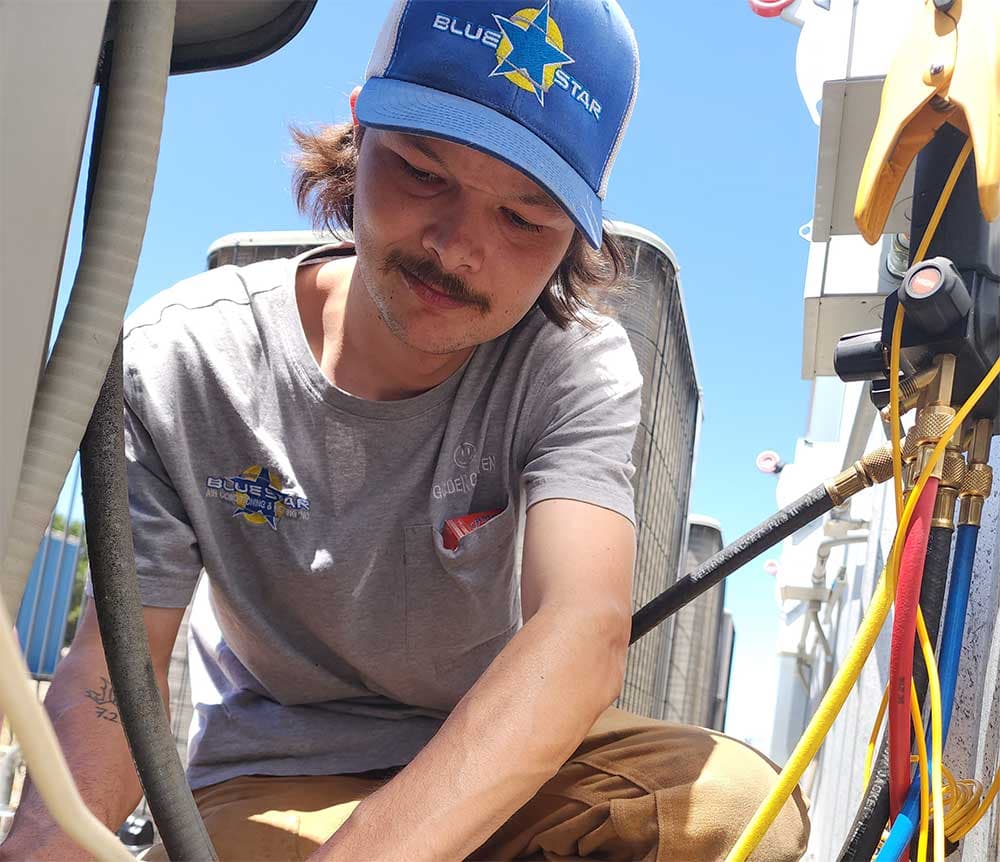 Blue Star technician in a Blue Star cap working among refrigerant gauge hoses at an outdoor AC unit