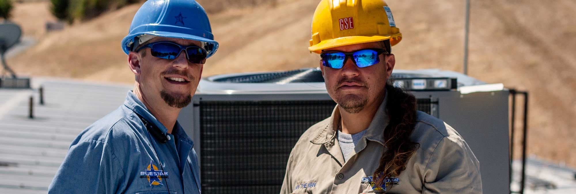 Two Blue Star technicians in hard hats and sunglasses on a roof in front of a condensing unit, with golden hills behind