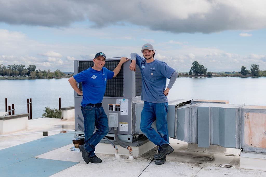 Two Blue Star technicians leaning on a newly installed rooftop unit with a calm waterway behind them
